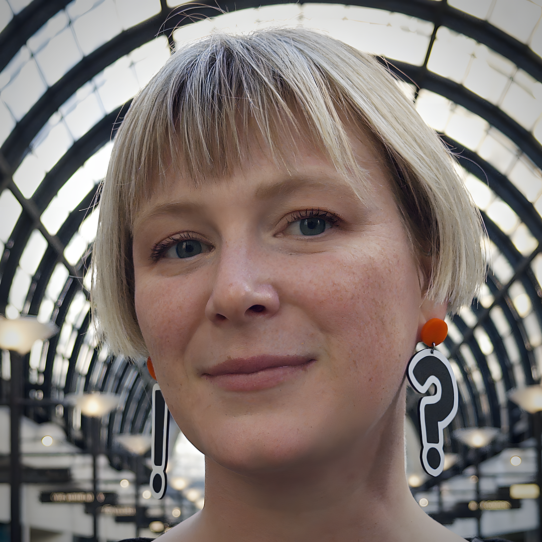 headshot of Katie Leavens in a blonde bob wearing punctuation earrings in front of an arch of windows and black metal framing